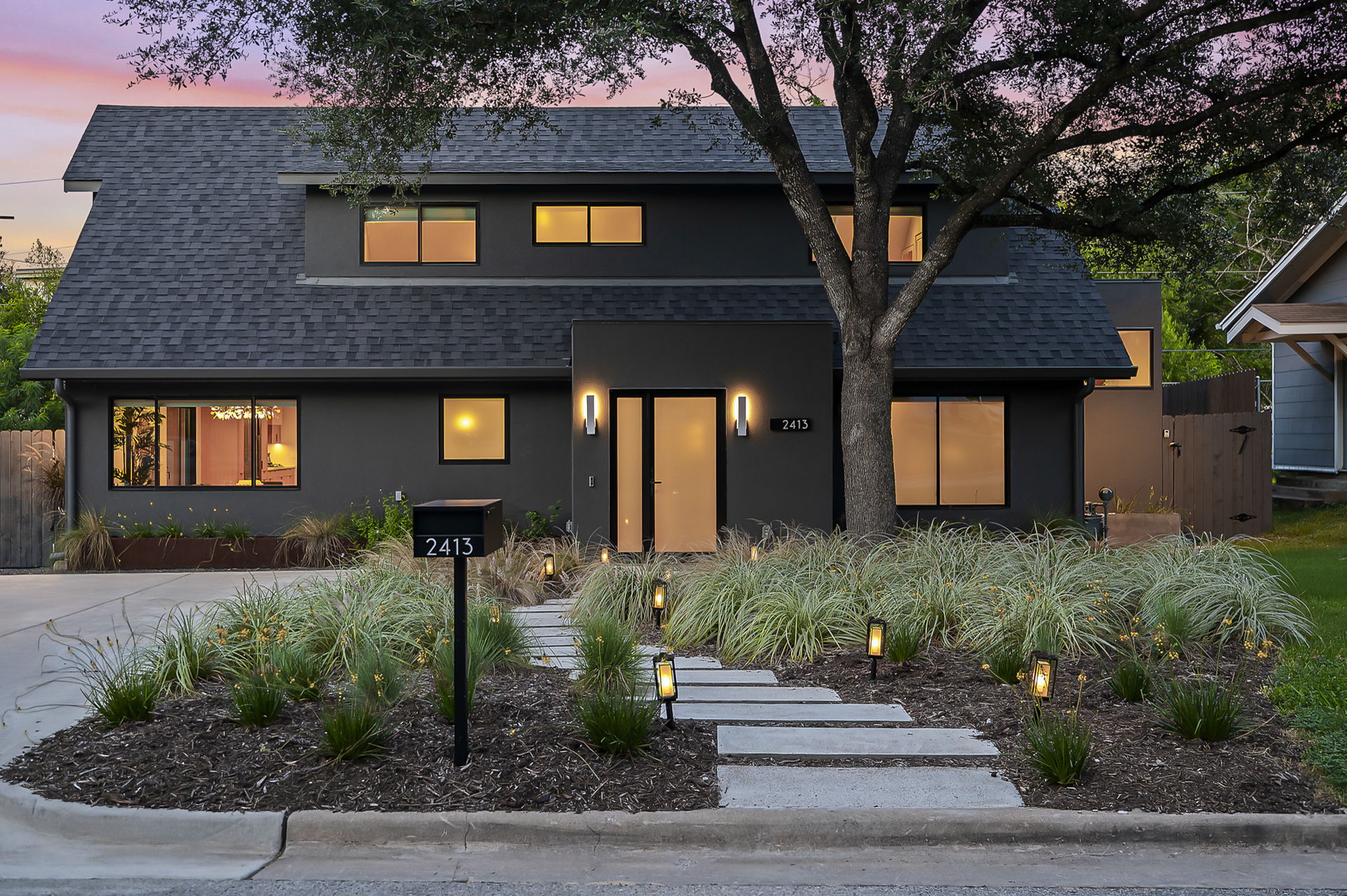 A contemporary two-story home with a dark gray facade, a minimalist front yard with a stepping-stone path leading to the front door, and subtle landscape lighting, all captured during a vibrant sunset.