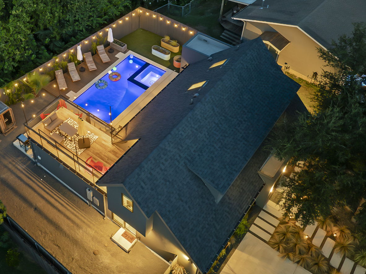 An aerial nighttime view of a modern home featuring an inviting backyard with a swimming pool, and a multi-level deck for lounging and entertaining, all illuminated with warm lighting.