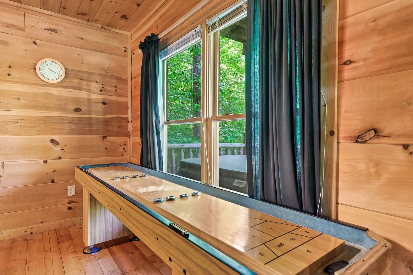 This rustic game area includes a shuffleboard table set against warm wood-paneled walls, with large windows framed by curtains that open to a lush green forest outside.
