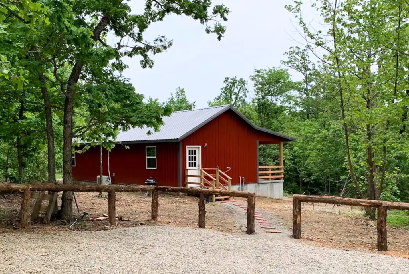 Misty Oak Lodge sits quietly among the trees with rustic fencing, a gravel drive, and a warm red exterior. The covered porch and wooded surroundings make it the perfect private escape for peaceful mornings, evening grilling.