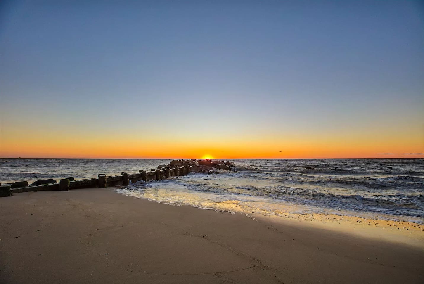 Capture a serene moment on the beach as the sun sets, painting the sky in warm shades of orange and gold. The gentle waves wash over the sand near a weathered wooden breakwater, offering a peaceful backdrop for your coastal getaway.