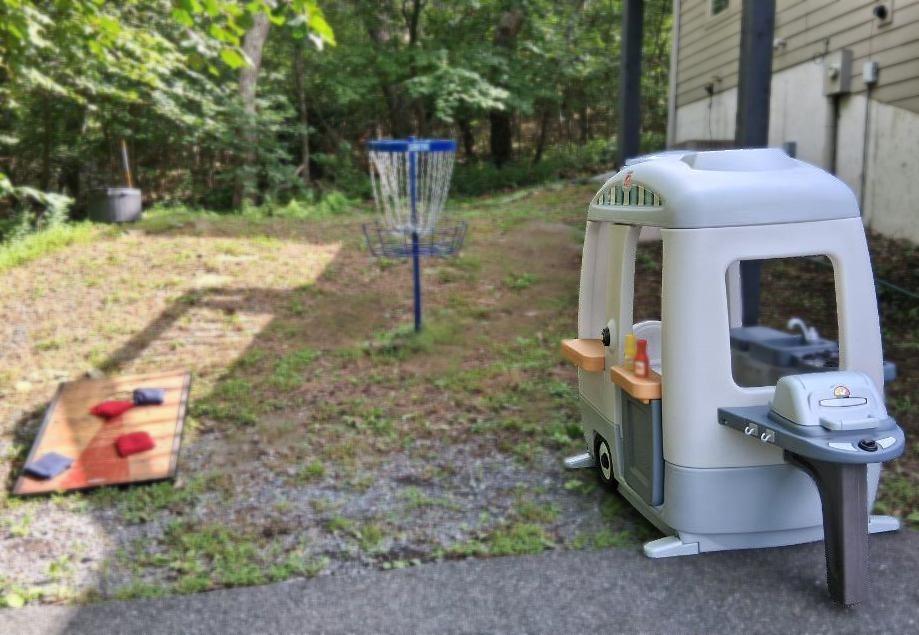 An open, casual play space with cornhole, a disc golf basket, and a toddler camper grill station—simple outdoor fun for all ages