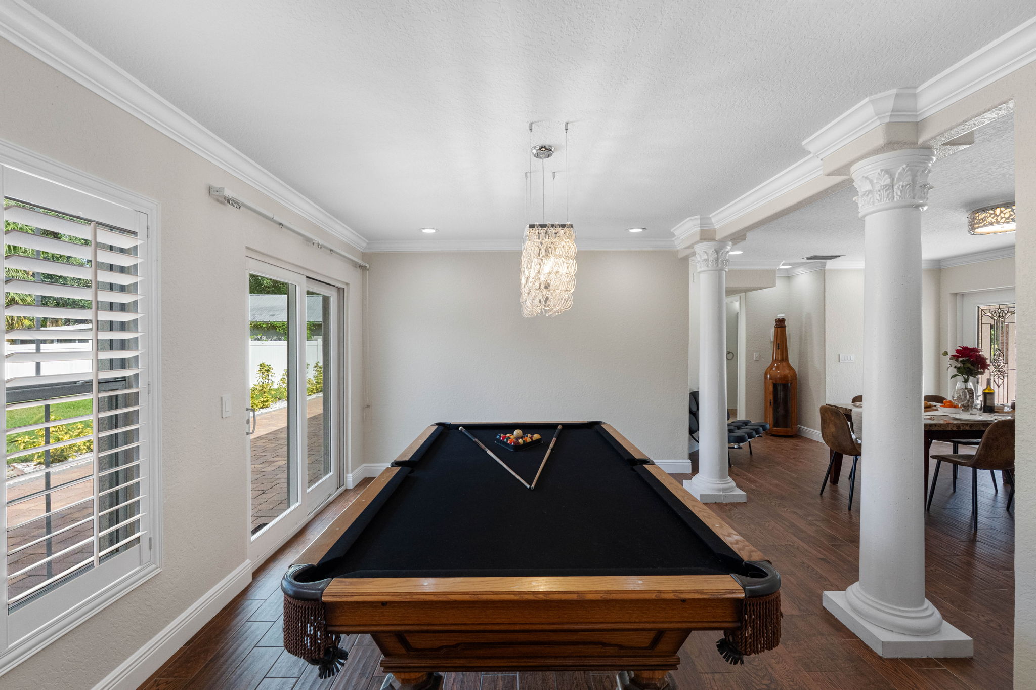 This elegant game room features a classic wood-framed pool table with a sleek black felt top, set beneath a statement crystal chandelier. Sliding glass doors open to a lush backyard with palm trees and serene water views.
