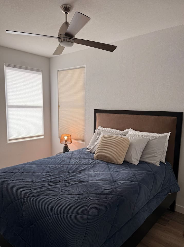 Queen bedroom with ceiling fan, closet storage, side table, and natural light from the corner windows.