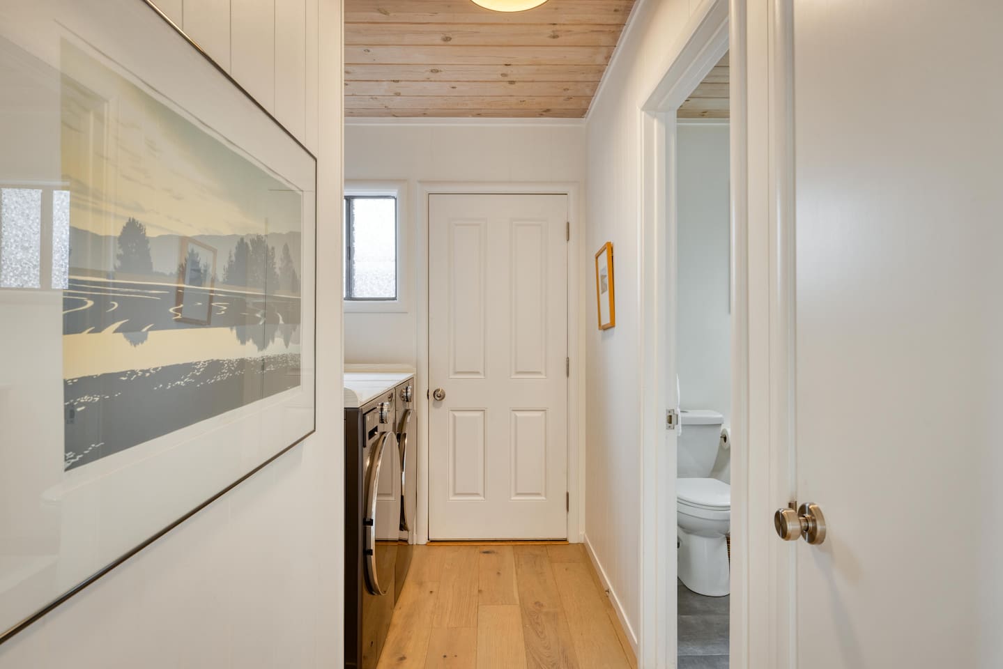 - Hallway with warm wood flooring and ceiling leading to laundry and bath  
- Side window allows natural light to brighten the functional corridor  
- Minimal wall art adds charm to this simple yet practical connecting space  
