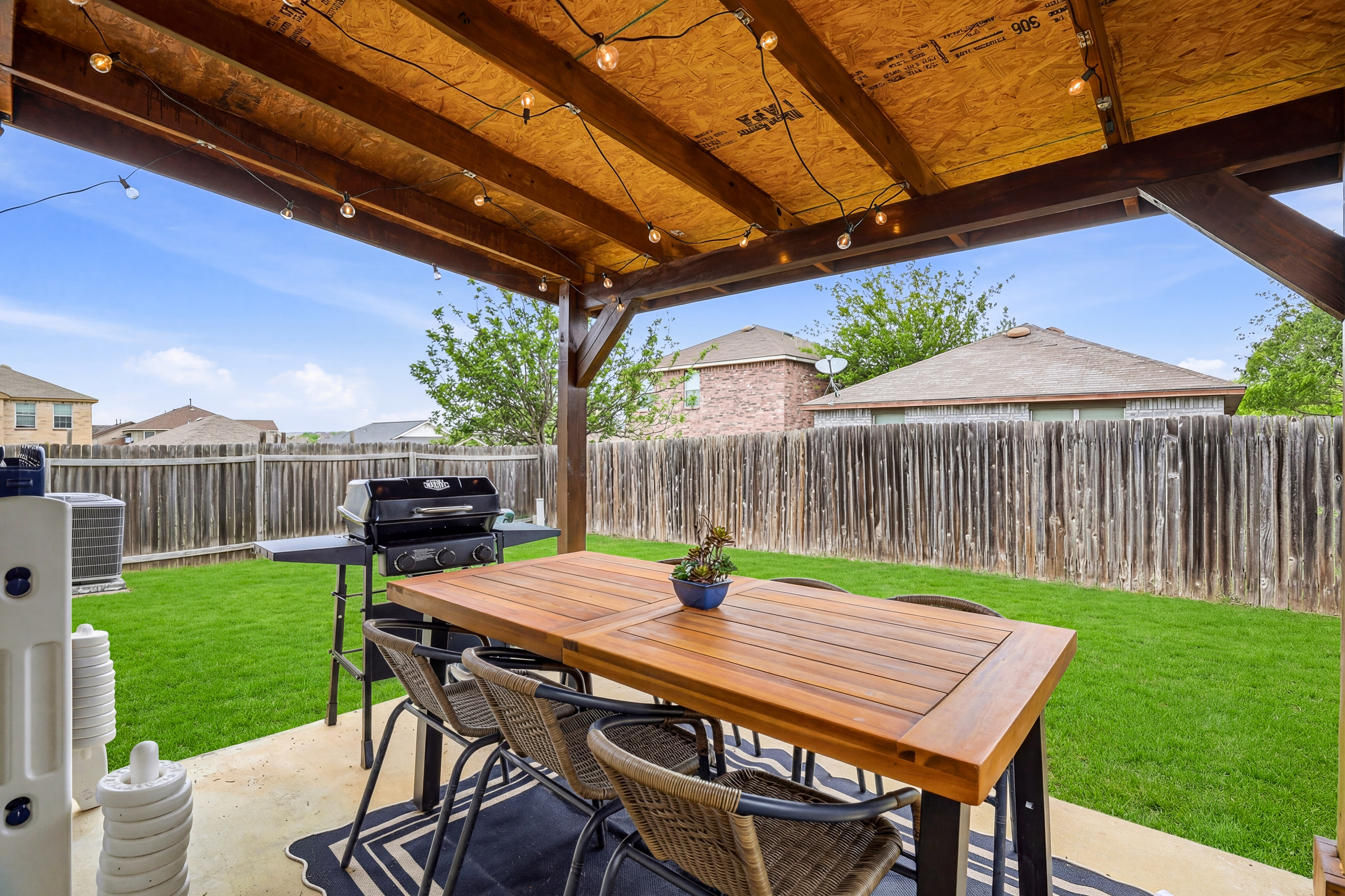 Covered backyard patio with wood dining table, string lights, and propane BBQ grill — perfect for alfresco meals and evening cookouts. Fully fenced for privacy with direct access from the kitchen.