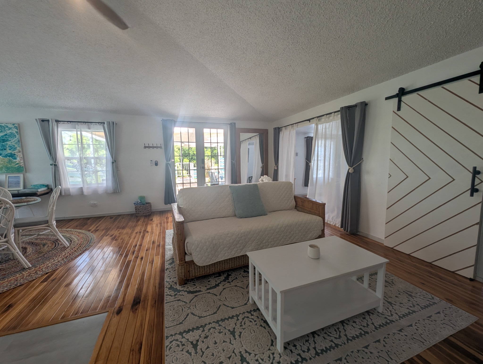 Upstairs living room at Villa Coral featuring a white couch, matching coffee table, patterned area rug, chevron barn door, and glass French doors leading out to a private rooftop patio in Dorado, Puerto Rico.
