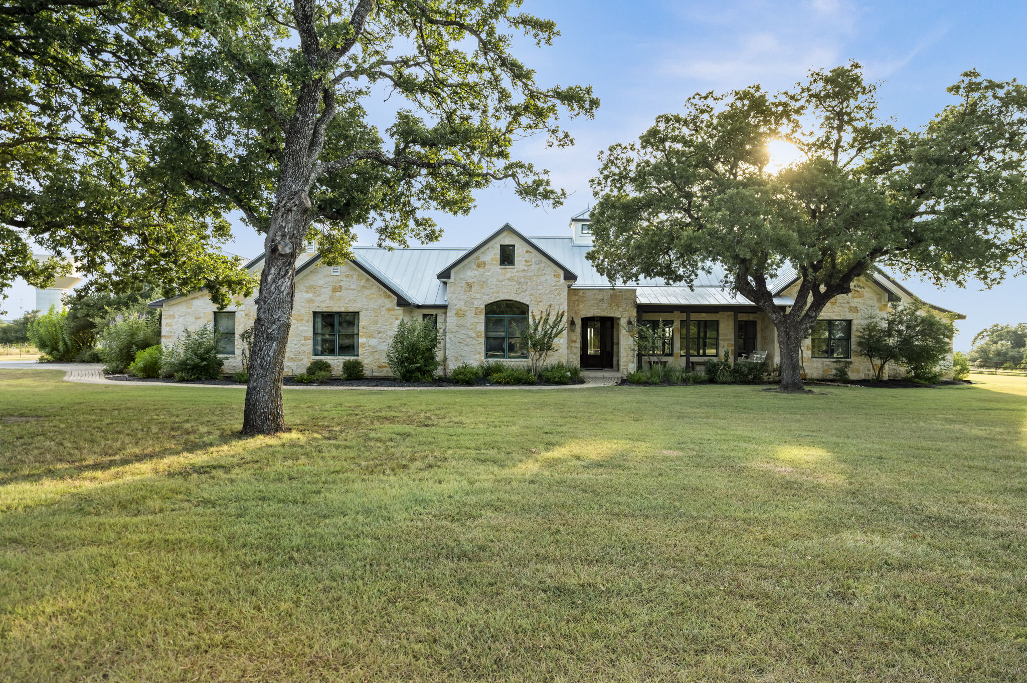 A stone exterior, expansive green lawn, and majestic oak trees under a clear sky.