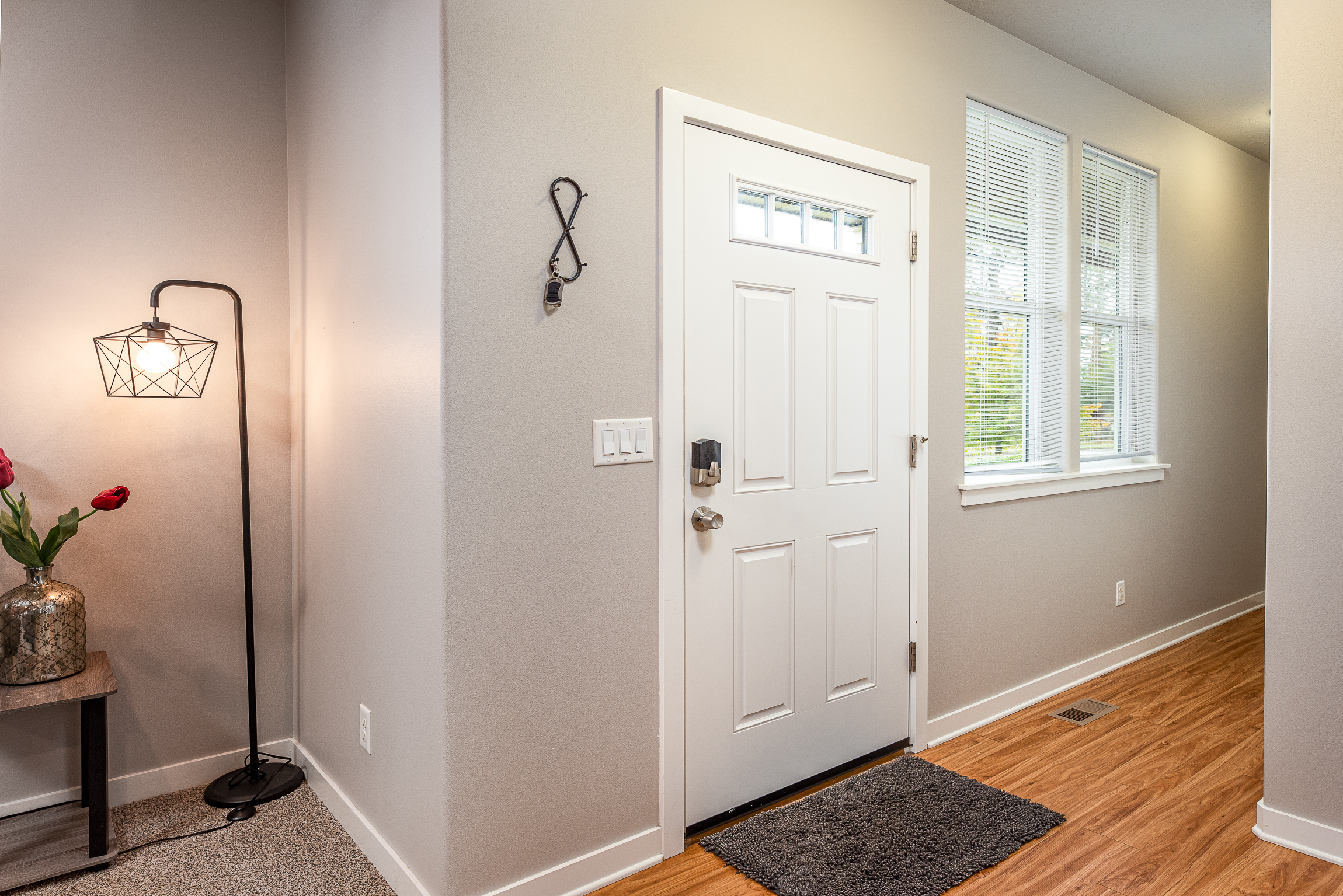 A well-lit entryway features a white front door with a small glass window, flanked by two vertical windows with blinds, while a stylish floor lamp with a geometric shade stands beside a table adorned with a vase containing red flowers, and a key holder is mounted on the wall.