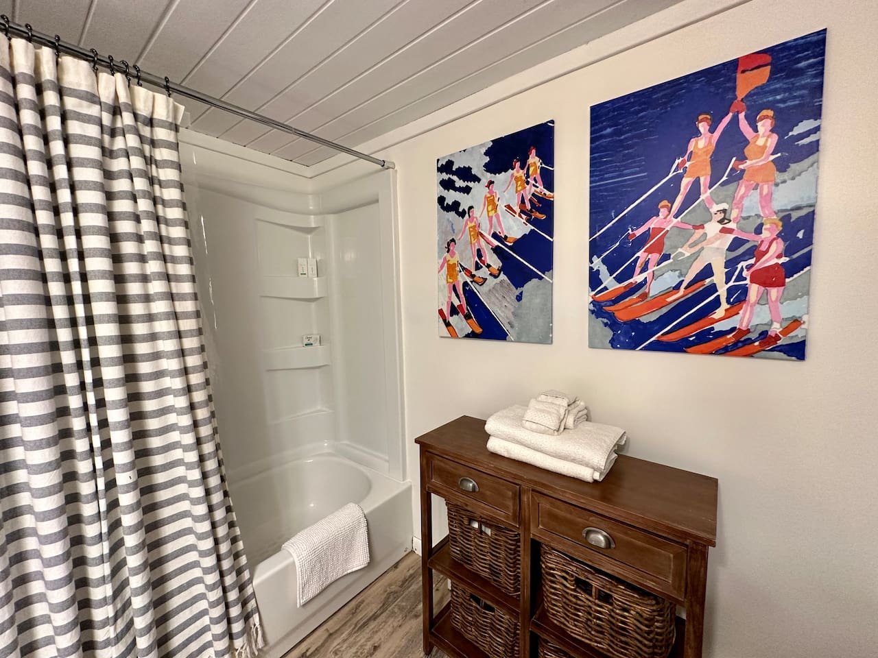 Fresh, coastal-inspired bathroom with a full tub/shower, striped curtain, soft towels, and fun vintage waterski art. A wood cabinet with woven baskets adds charm and storage, making this space both functional and inviting for you.