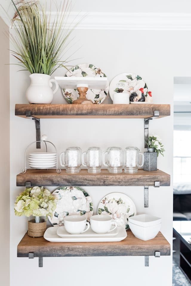 These charming rustic floating shelves are fully stocked with essentials, including mason jar mugs, decorative floral plates, and clean white bowls. Everything is neatly organized and easily accessible for your dining needs.