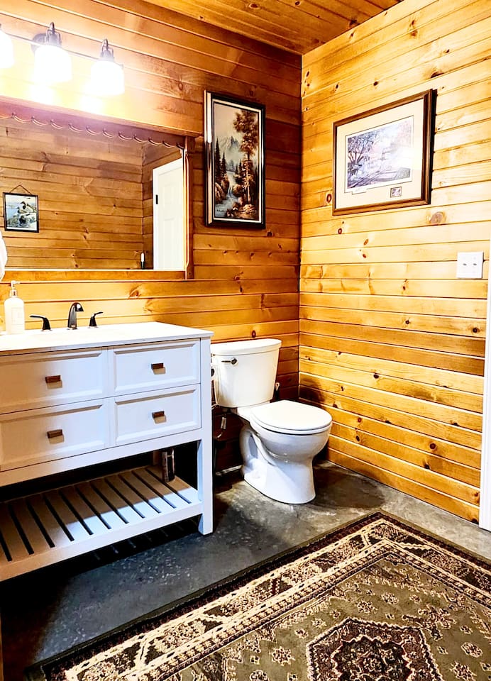 This charming rustic bathroom features warm cedar walls, a modern white vanity with black hardware, soft lighting, and classic decor. A cozy rug and natural textures create a clean, welcoming space to refresh and unwind after a day outdoors.