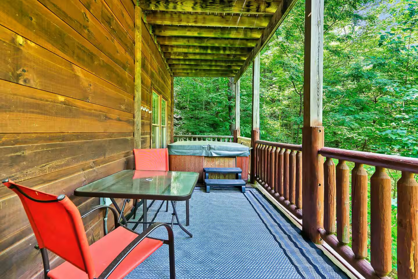 This inviting cabin deck includes a bubbling hot tub, surrounded by a rustic wooden railing and nestled in a serene forest setting, perfect for unwinding in nature.