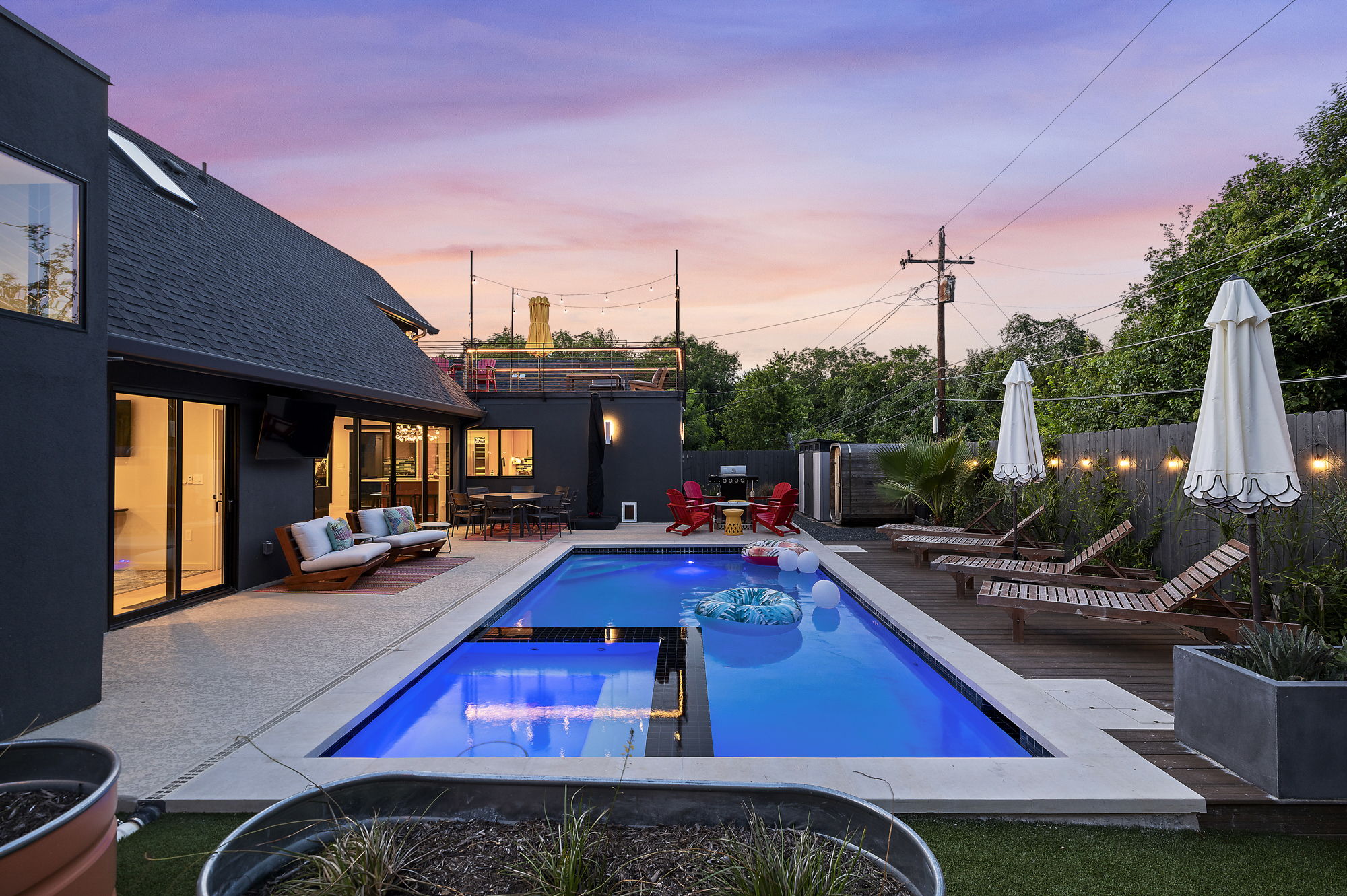 A luxurious backyard at sunset featuring a sparkling swimming pool, a spacious deck with multiple lounge chairs and a dining area, all set against a modern dark gray home with large windows and a rooftop deck.