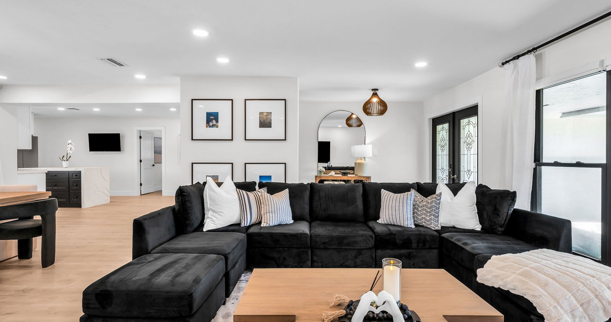 Elegant open-concept living room featuring a plush black velvet sectional, 75” smart TV, and designer mid-century modern furnishings. Floor-to-ceiling windows and French doors fill the space with natural light and connect seamlessly to the outdoor pool.