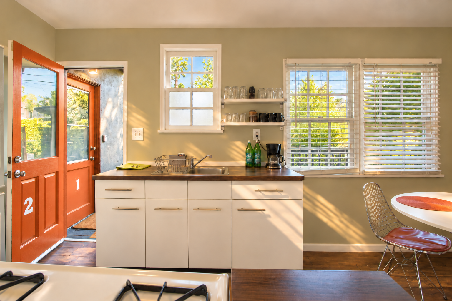 Another kitchen perspective shows white cabinets, the wood countertop and stainless-steel sink beneath the windows. Open shelves keep glassware close at hand, with the round dining table to the right and the orange exterior doors visible to the left.