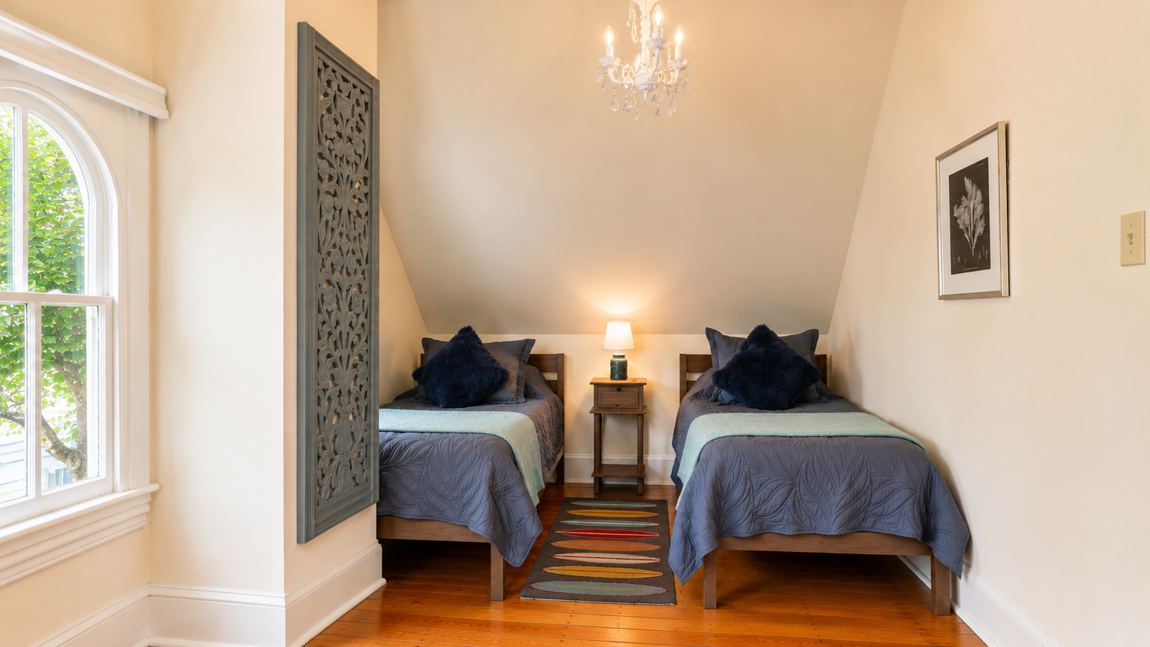 The third floor twin bedroom — tucked under the eaves with a crystal chandelier, original hardwood floors, and arched windows that look out into the treetops.