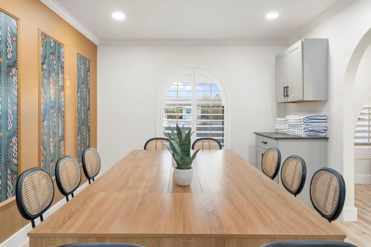 Gather the whole group around this oversized table, built for long dinners and even longer conversations. Woven-back chairs and vibrant wall art add warmth, while the sunlit archway window keeps the space feeling open all day.