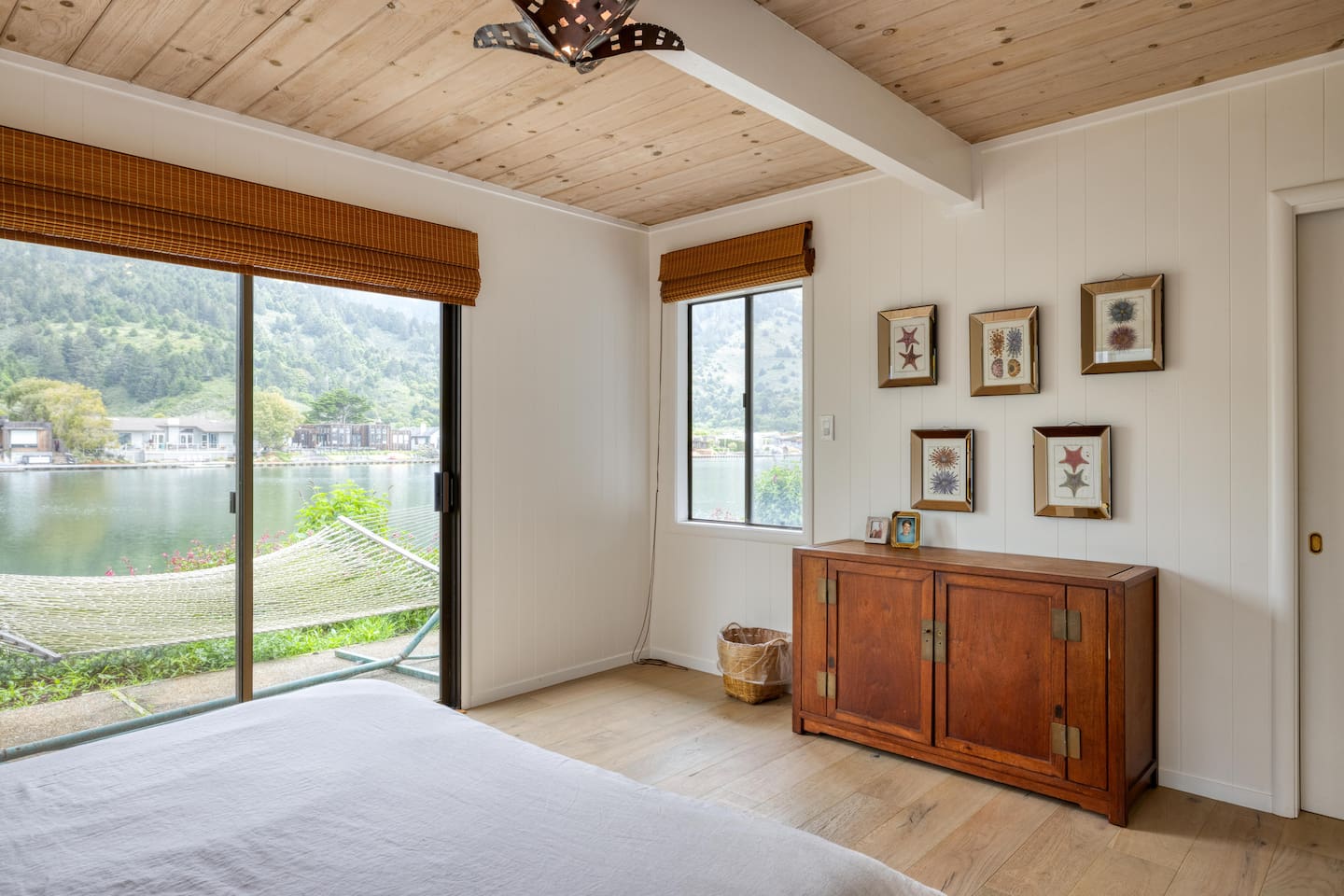 - Bedroom with queen bed, wood ceiling, and direct patio access with hammock  
- Framed artwork and a wooden dresser add charm to the cozy sleeping space  
- Windows on two sides provide natural light and water views from the bed  
