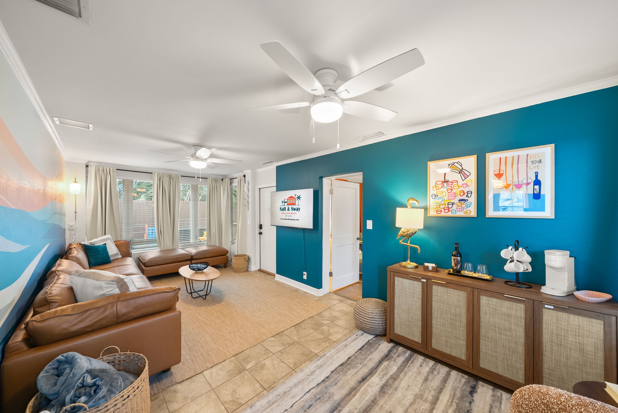 The wide-angle living room view shows how the coastal design
layers together. The leather sectional anchors the seating, the
surf-wave mural fills the wall, and the backyard is right out the door.