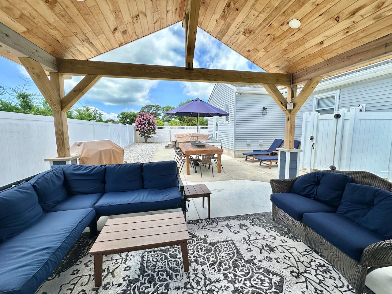 Relax under this wooden pavilion featuring navy sectional seating and a patterned rug. The space offers views of the hot tub, outdoor dining area, and grill, providing a perfect setting for all-day entertainment.