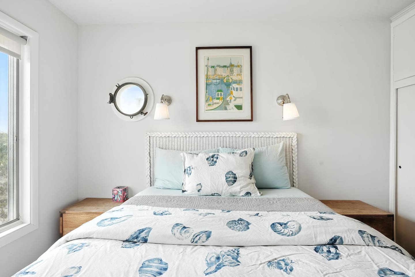 - Bright coastal themed bedroom with white headboard and sea inspired bedding  
- Features porthole style window, wall sconces, and framed art  
- Crisp white walls and light wood furniture enhance the airy atmosphere