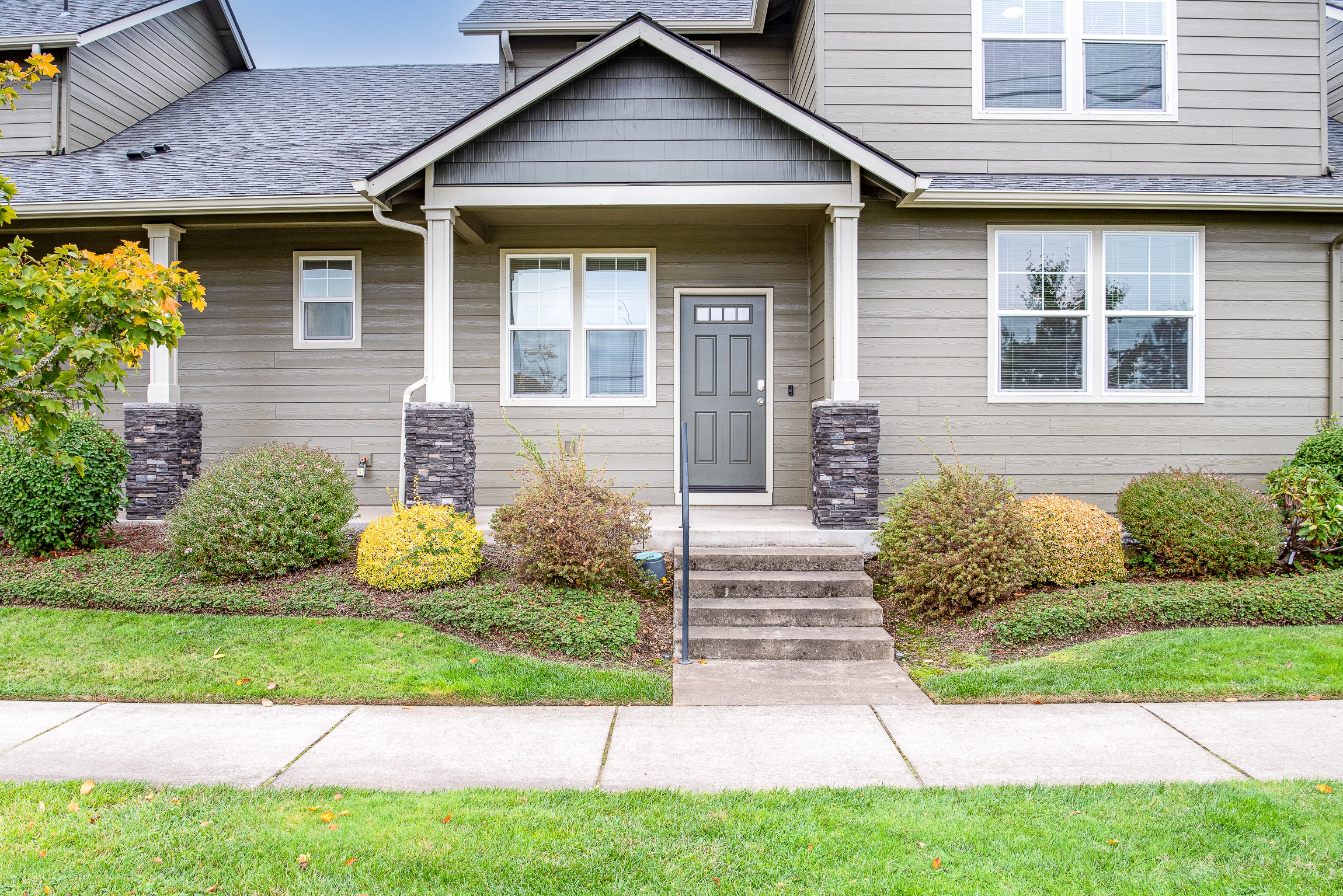 The image shows a well-maintained, two-story house with a gray exterior, featuring stone pillars flanking a dark front door, a small porch with steps, and landscaped bushes and plants on either side, set against a clear sky.
