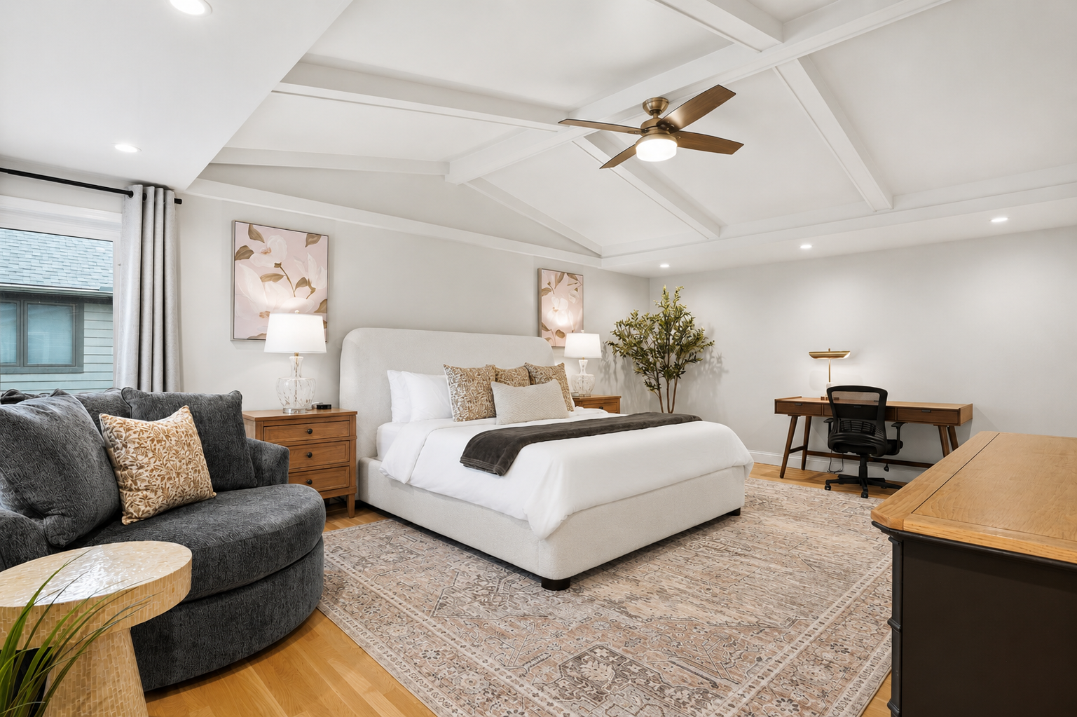 Relax in this spacious primary suite featuring a plush bed, vaulted ceiling with modern fan, natural light, and a comfortable work and seating area designed for both rest and productivity.