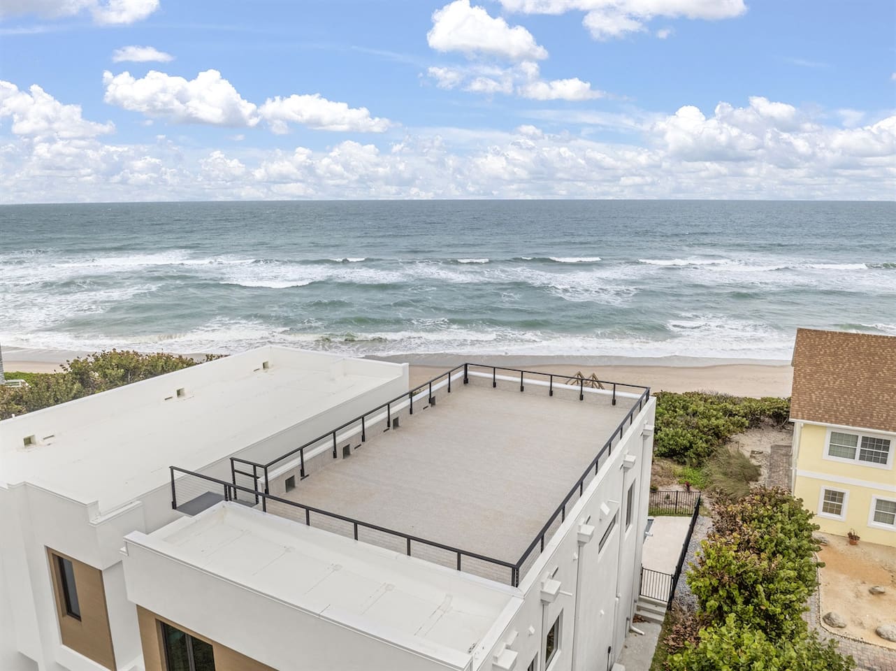 Captured by drone, the rooftop offers a stunning view of the neighborhood's houses, with the ocean stretching beyond and lush greenery surrounding the area, creating a peaceful and scenic escape.