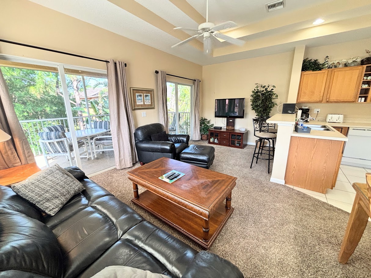Comfortable living area with high ceilings, tile flooring, cozy seating, and two sets of sliding glass doors that bring in natural light and lead out to the private screened balcony.