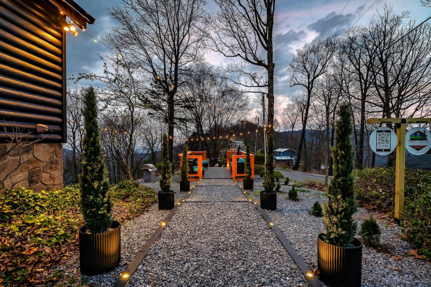 An inviting pathway lined with manicured potted trees leading to a scenic hangout deck, BBQ, outdoor game, and firepit area - perfect for relaxing, socializing, and enjoying mountain views.