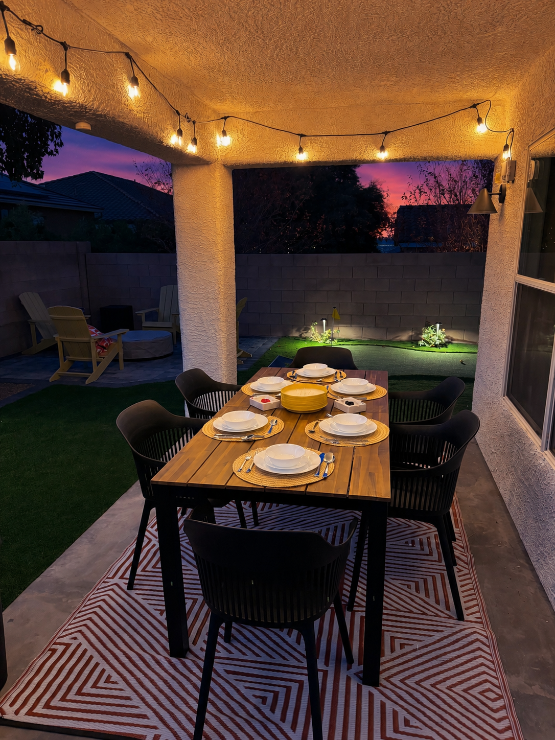Covered outdoor 6-person dining area with soft string lighting and our firepit beyond, at night. -Adventure Awaits.
