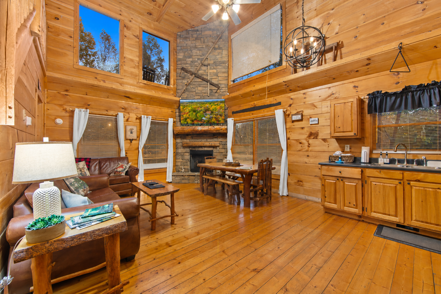 This open-concept cabin living area boasts cozy leather couches, a rustic wooden staircase, and a fully equipped kitchen with black appliances, all set within a warm, wood-paneled interior.