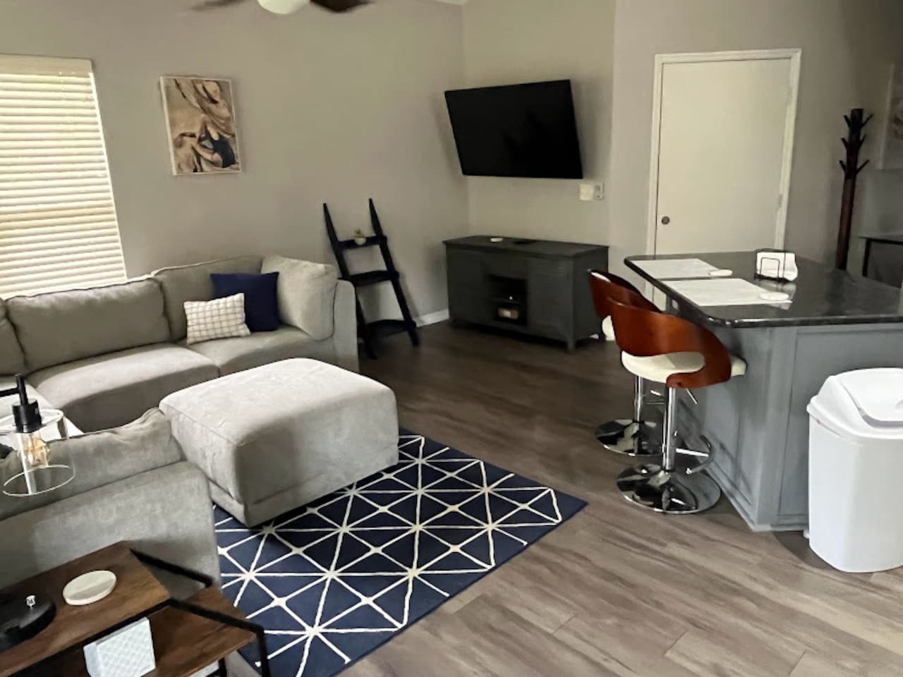 Comfortable living area with modern seating, wood-look flooring, wall-mounted TV, and an open view toward the kitchen.