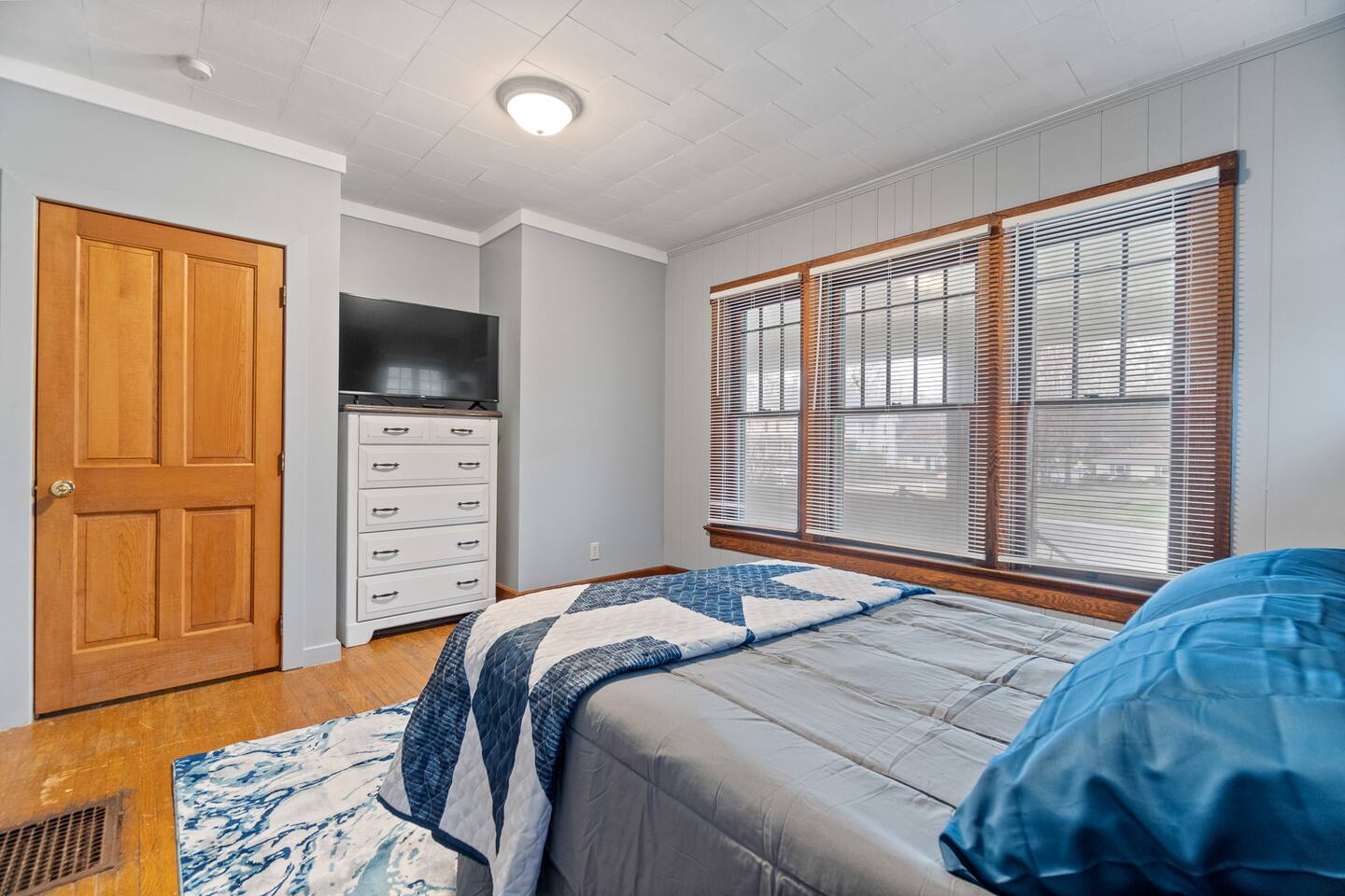 "Charming bedroom featuring a modern sofa with pillows, large windows next to the bed, a sleek cabinet, widescreen TV mounted above, and a door leading to the bathroom—designed for comfort, relaxation, and modern convenience.