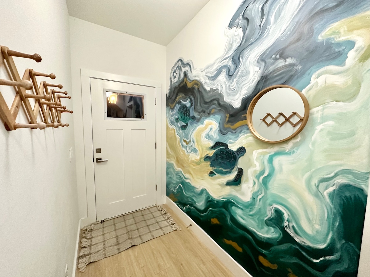 You come in through this. Hooks for wet towels on one side, a hand-painted sea turtle riding the surf on the other, and the whole house opening up beyond it. Guests photograph this wall before they unpack.