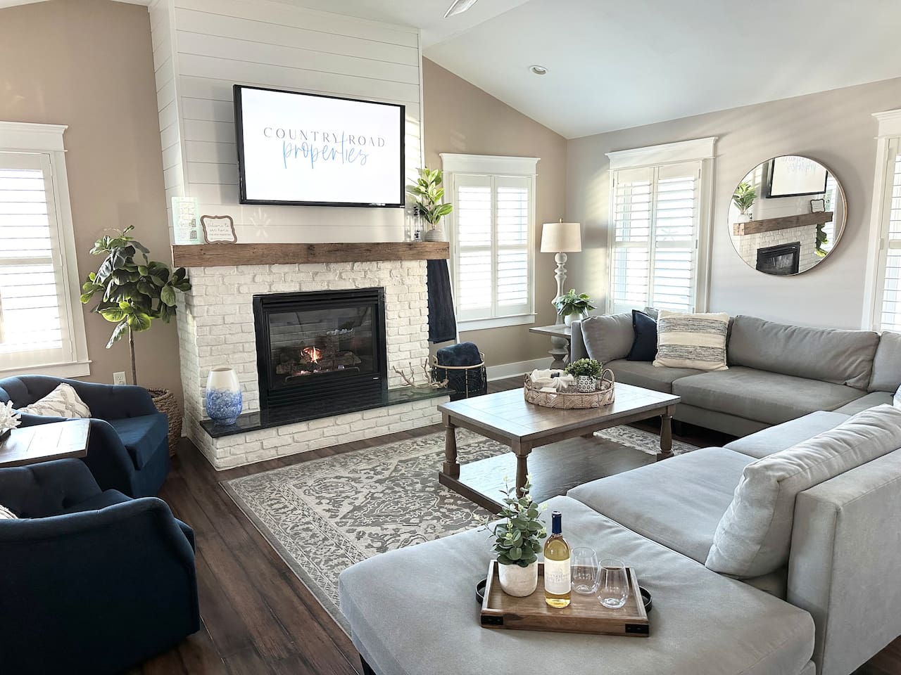 Gather in this sun-drenched living room featuring vaulted ceilings and a cozy white brick fireplace. The sprawling sectional and navy accent chairs offer plenty of space to relax and watch a movie after a long day at the beach.