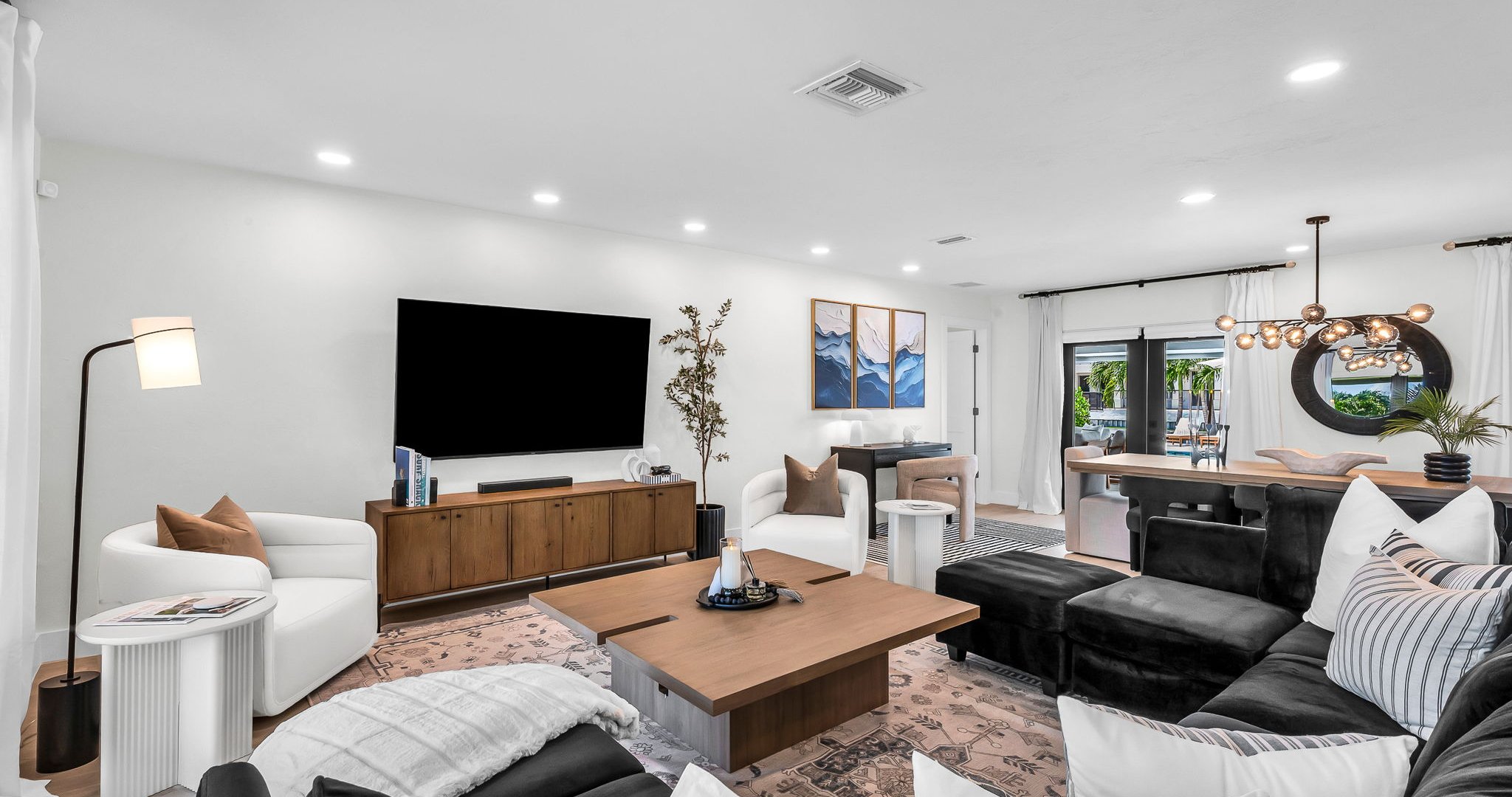 Elegant open-concept living room featuring a plush black velvet sectional, 75” smart TV, and designer mid-century modern furnishings. Floor-to-ceiling windows and French doors fill the space with natural light and connect seamlessly to the outdoor pool.