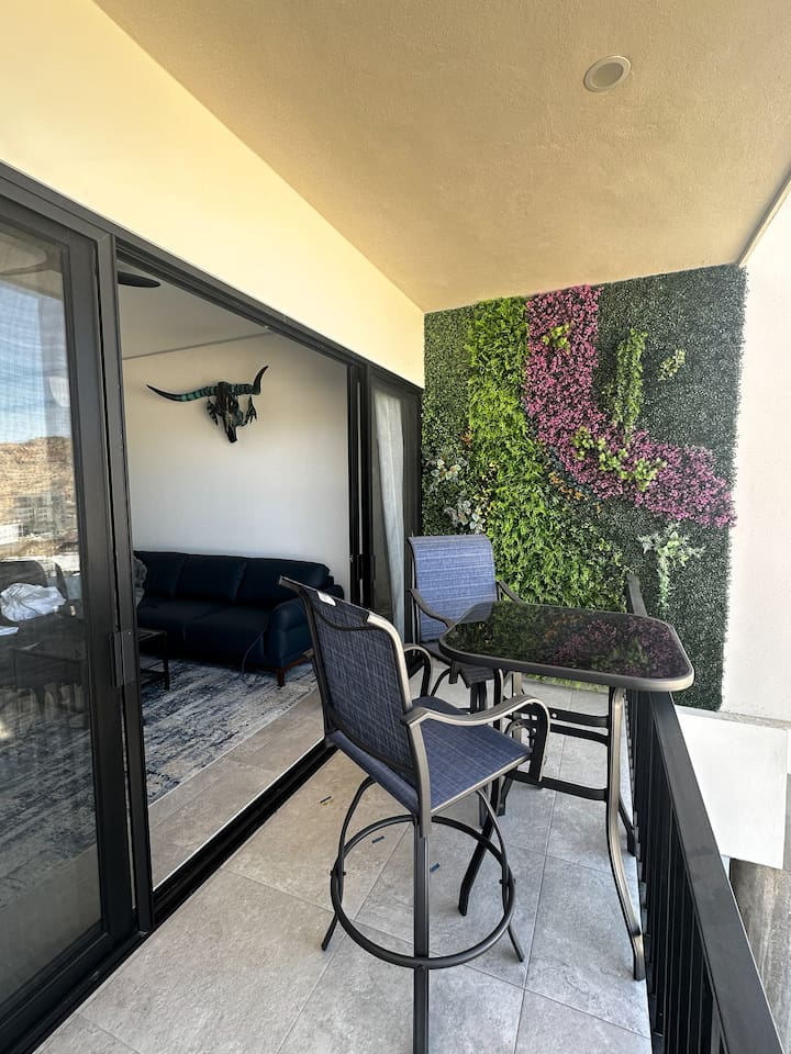 Step from the cozy living room to the balcony, featuring a stylish high-top outdoor dining table and a serene outdoor nature wall.