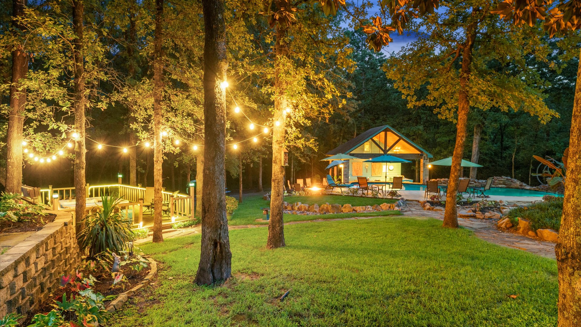 A dreamy nighttime retreat! This beautifully lit backyard features a sparkling pool, cozy fire pit, and a charming outdoor kitchen. Relax under the umbrellas or take a dip—perfect for unwinding in a serene setting.