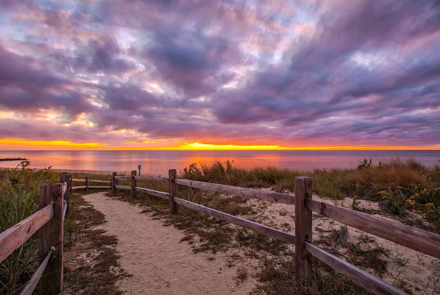 End your day with a breathtaking sunset view over the water. This scenic sandy path, lined with rustic wooden fencing, leads you directly to the beach to enjoy vibrant purple and orange skies as the sun dips below the horizon.