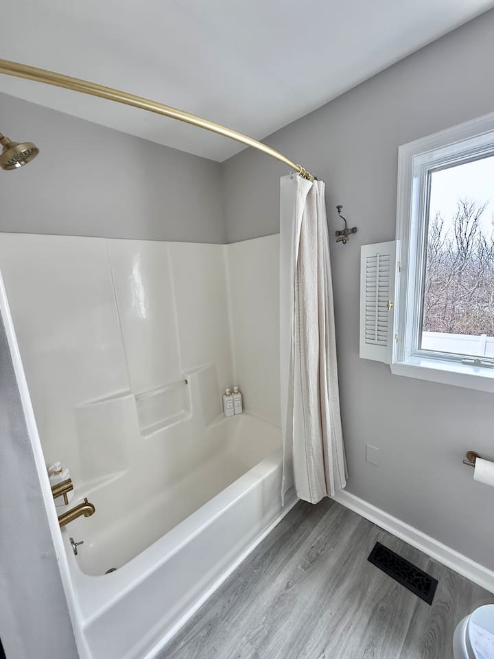You will see a clean, modern bathroom space featuring a white tub-shower combination accented with elegant gold-tone fixtures. A light, neutral-toned shower curtain and shuttered window complete the serene, coastal aesthetic.
