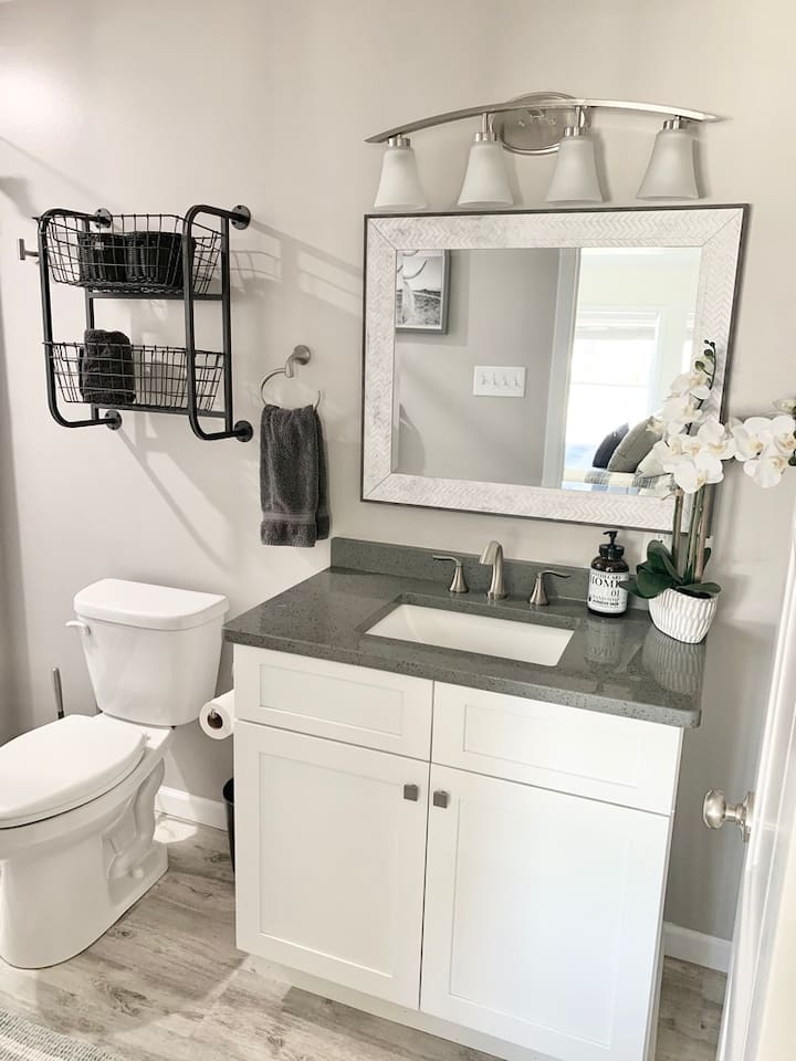 Enjoy a modern and clean guest bathroom featuring a sleek grey quartz countertop and a white vanity. The space is thoughtfully designed with industrial-style wall baskets for storage, a textured frame mirror, and elegant orchid accents.