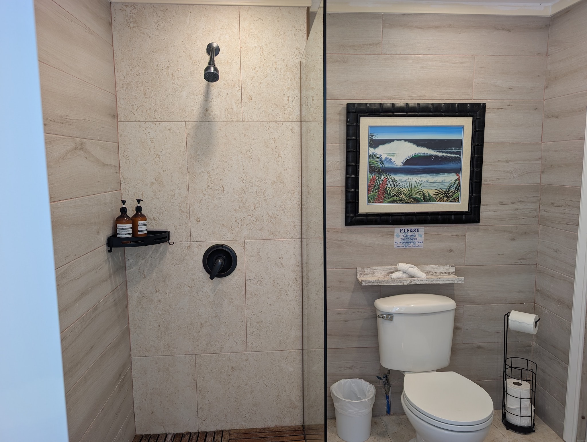 Poolside cabana bathroom at Villa Coral featuring a walk-in shower with black fixtures, wood-look tile accents, travertine walls, and ocean surf artwork in Dorado, Puerto Rico.
