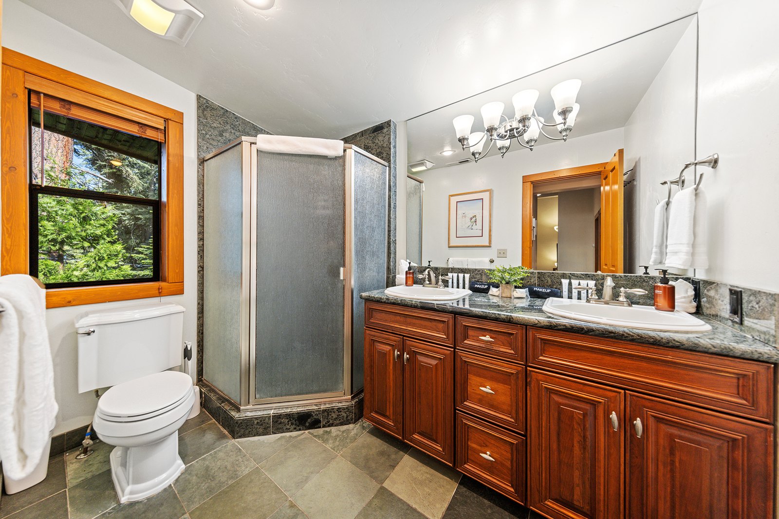 Rinse off the trail day in a double-vanity bath — corner shower, stone counters, and pines framed in the window.