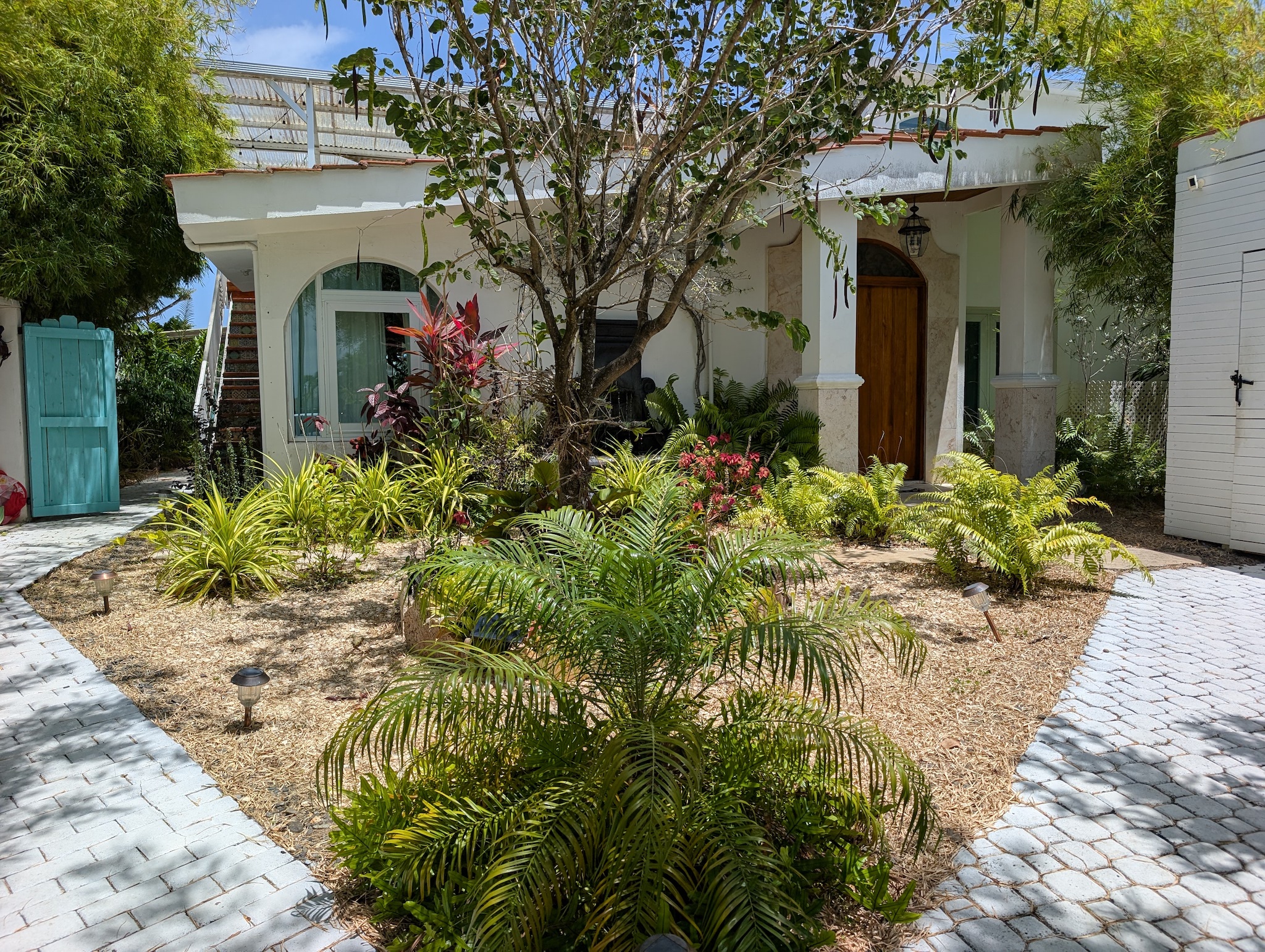 Wide shot of the tropical entry courtyard at Villa Coral featuring stone paver walkways, a palm tree garden, grand pillars, and a custom wood front entry door in Dorado, Puerto Rico.
