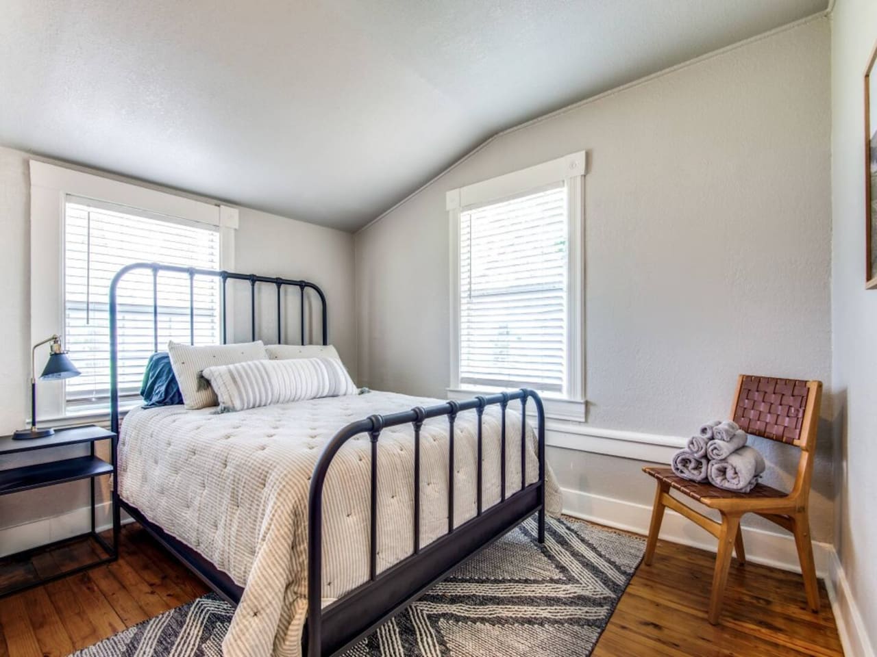 Light-filled queen bedroom with iron-frame headboard, woven rug, hardwood floors, and bright window. Sleeps 2 as part of a 3BR/2.5BA farmhouse sleeping up to 7 near The Milestone and The Grove wedding venues in Aubrey TX.