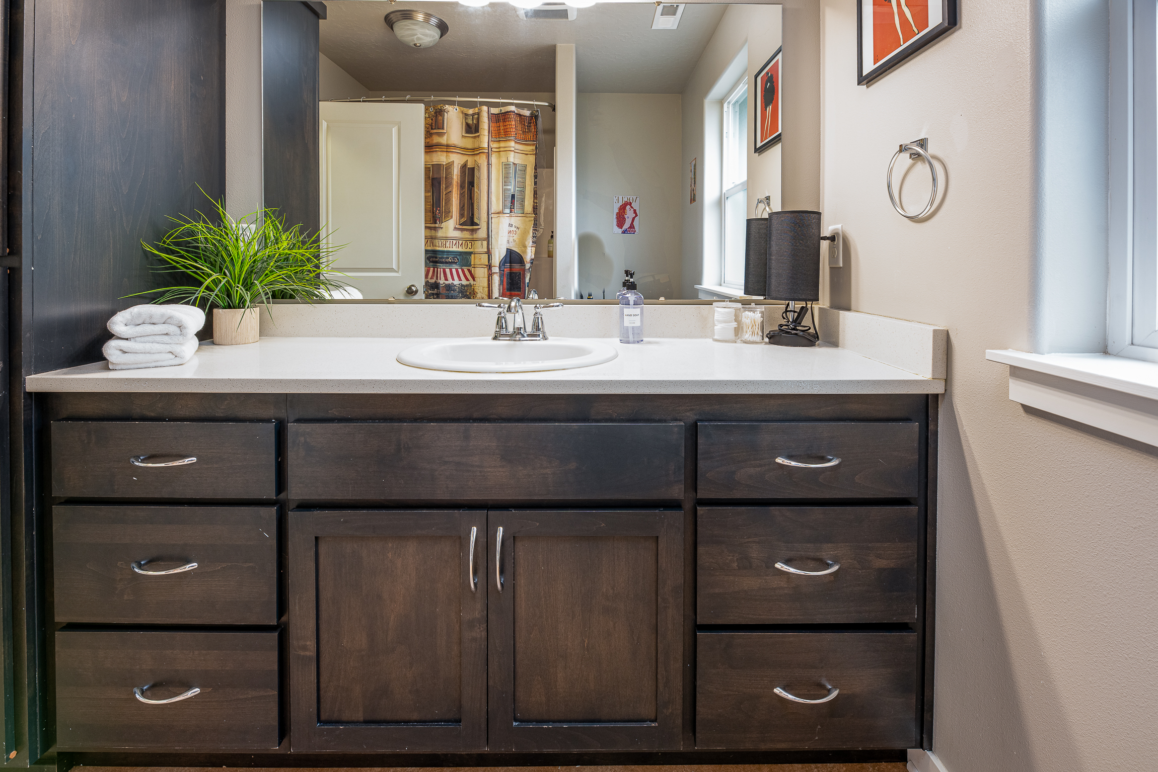 A modern bathroom vanity with a dark wood finish features a white countertop, a sink with chrome fixtures, neatly folded towels, a small potted green plant, a bottle of hand soap, and cotton swabs, all reflected in a large mirror above; behind, a decorative shower curtain with a cityscape design and a bright window provide additional context.