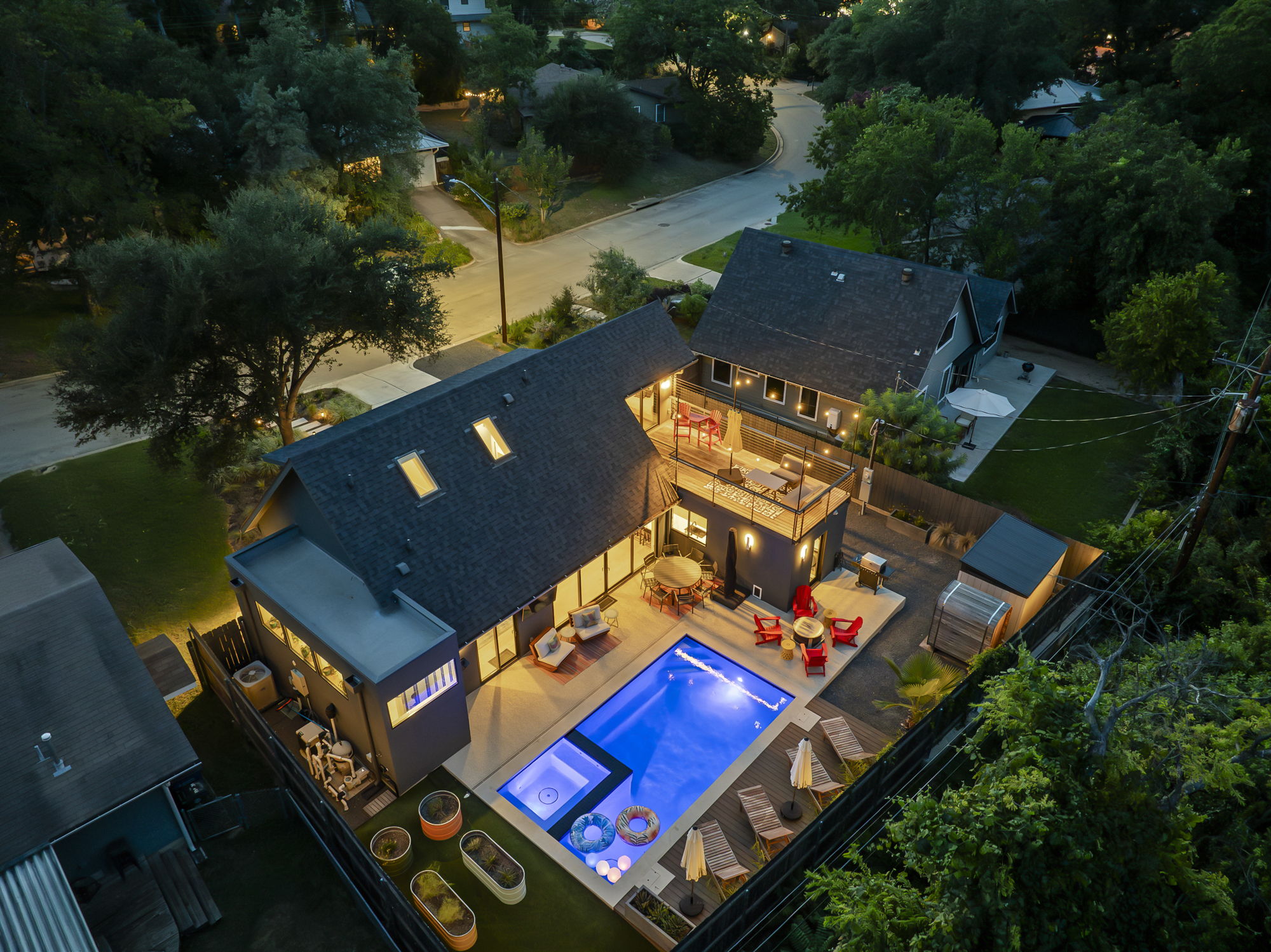 An aerial nighttime view of a modern home, showcasing a large swimming pool, a hot tub, and a multi-level deck for lounging and entertaining, all beautifully lit by string lights and outdoor fixtures.