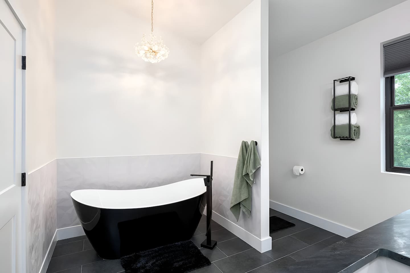 Indulge in a spa-like experience in this luxury master bath. It features a stunning freestanding black soaking tub, elegant crystal chandelier, and modern matte black hardware. A serene, high-end retreat designed for ultimate relaxation.